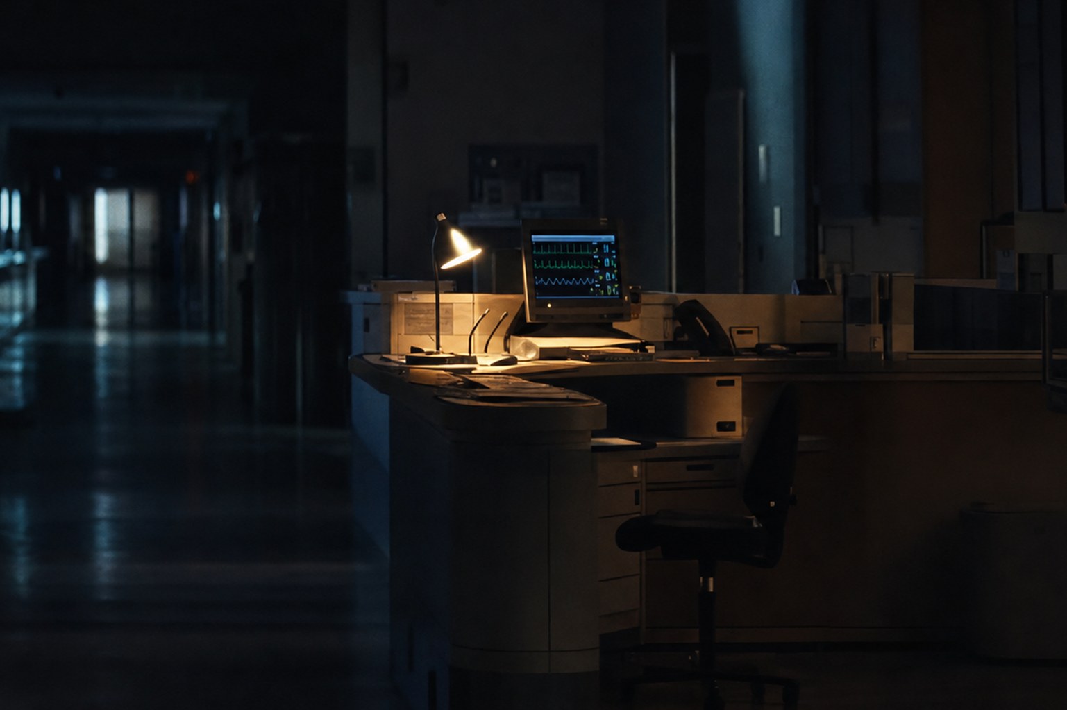 An empty hospital nurses station at night, lit by a single cool overhead panel, with a keyboard and stacked paper charts on the counter.