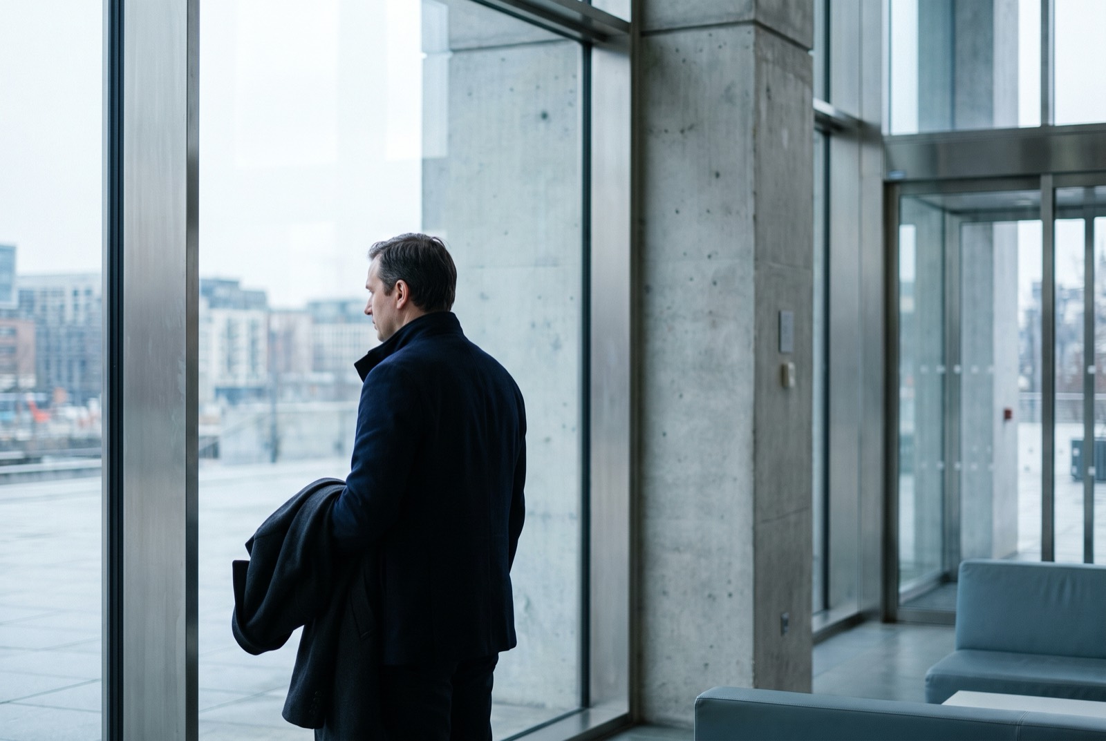 A man in his forties stands at a tall window in a quiet building, coat over his arm,
                      looking out at the city.