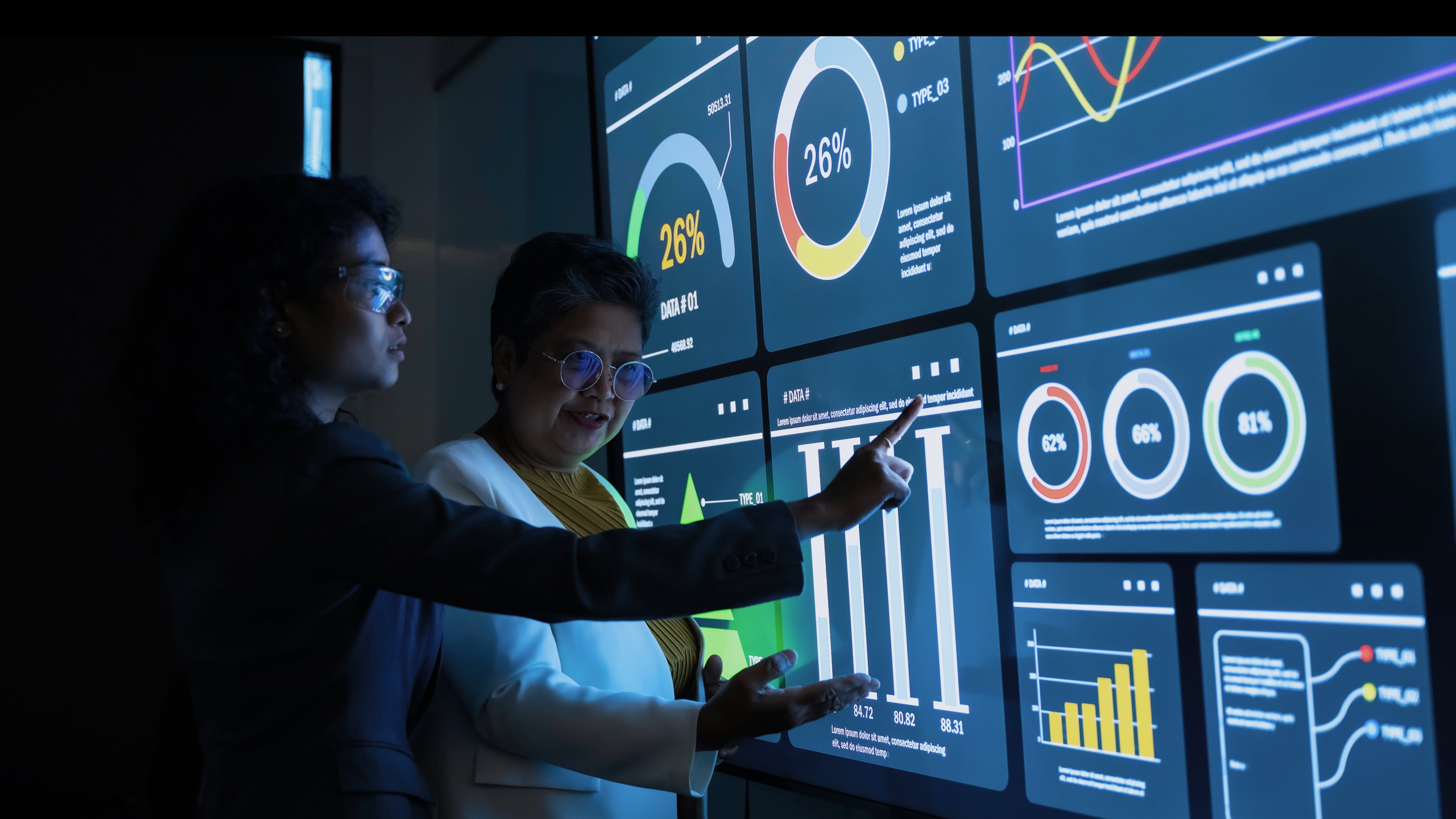 Two executives reading live analytics on a wall of dashboards in a darkened operations room.