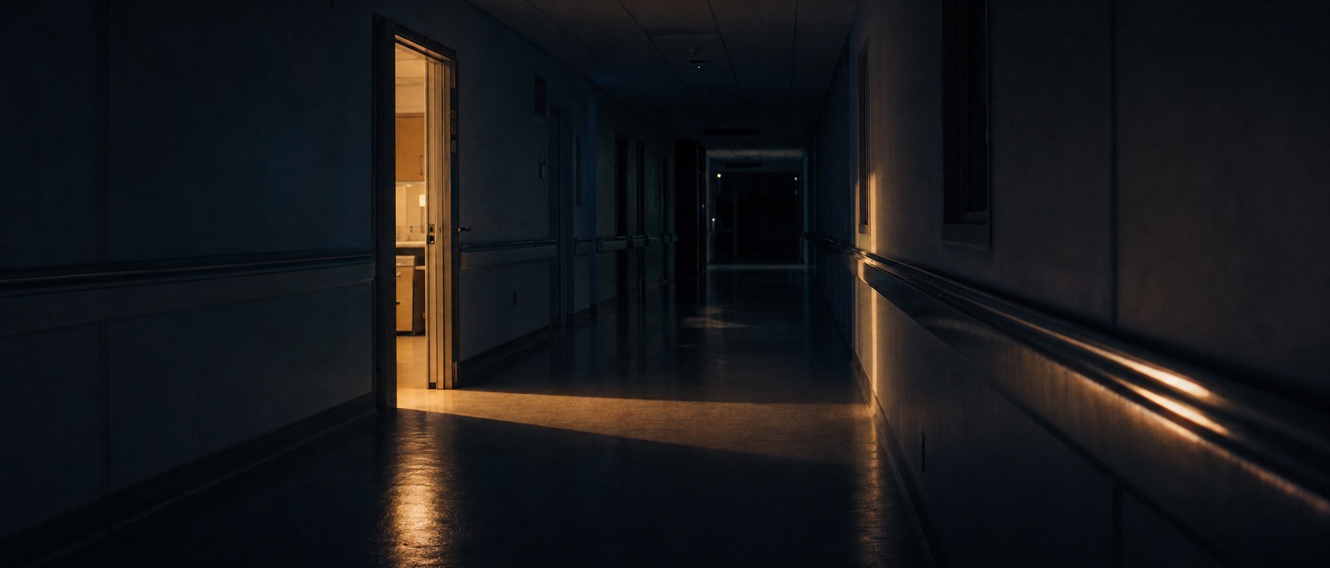 A darkened hospital corridor lit only at its far end, where a single distant figure stands between treatment room doors.
