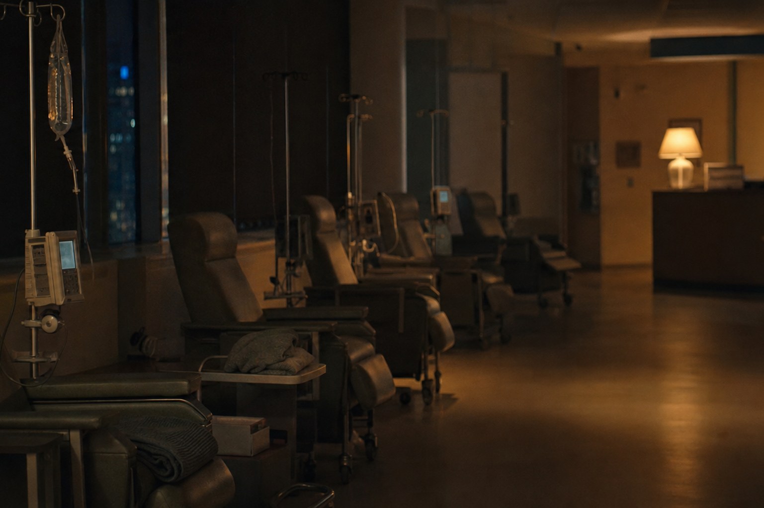 An empty clinical workstation at the end of a shift: a vacant chair, a coiled stethoscope and a cold paper cup lit by a single overhead panel, the rest of the room in darkness.