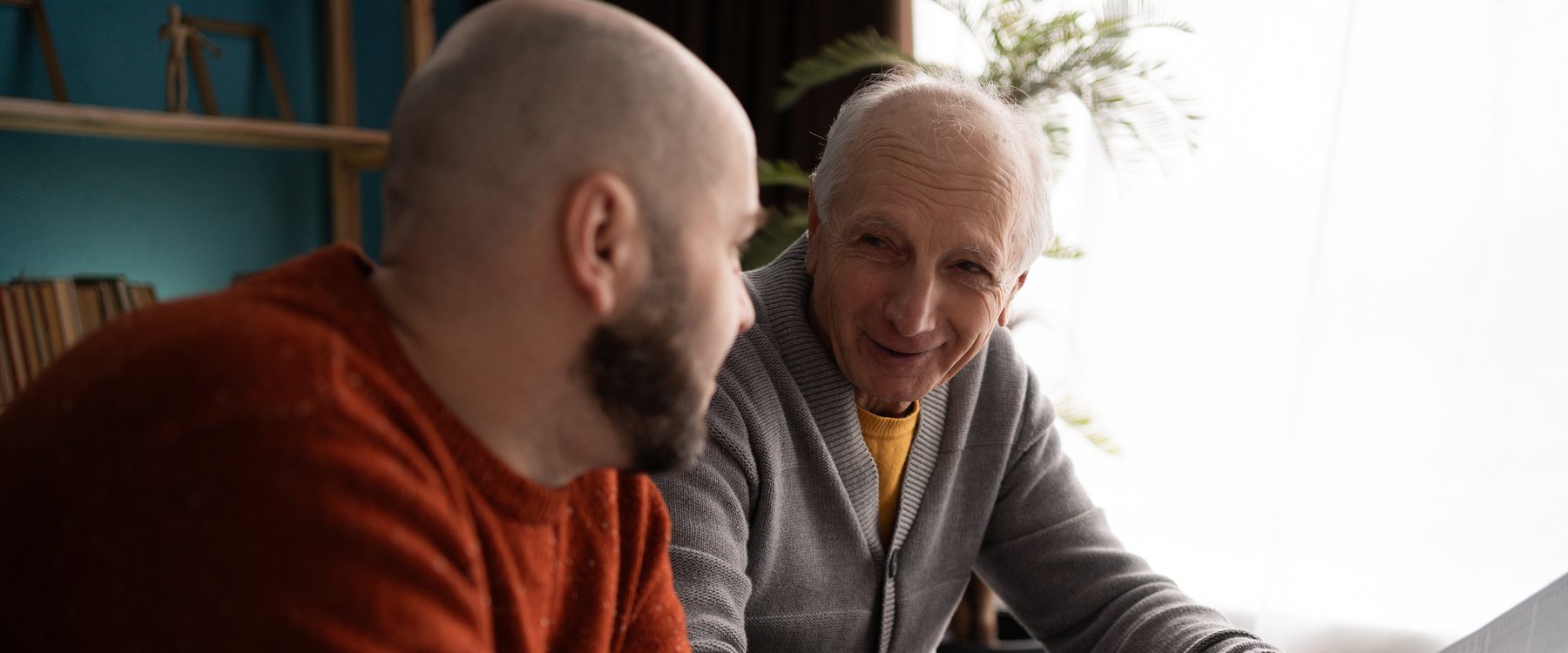 An adult son and his elderly father sitting together at a table, going through paperwork in warm conversation.