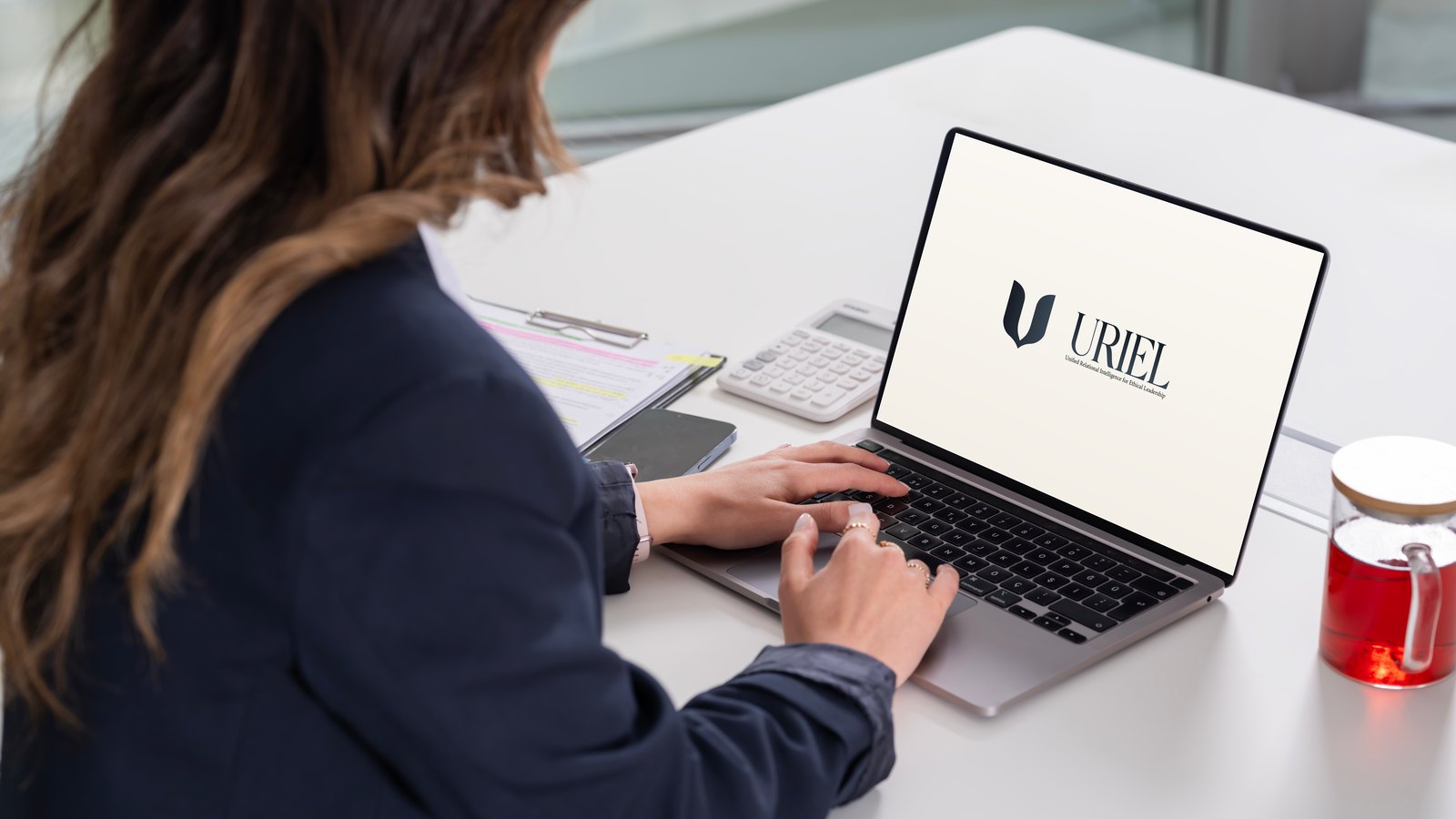 A woman at a desk in a bright office, working at a laptop whose screen shows the URIEL lockup.
